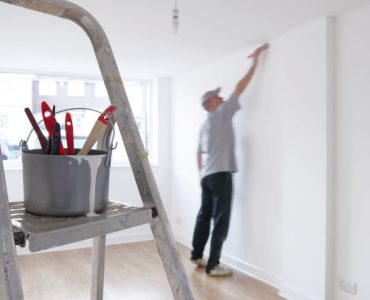 Une échelle et un pot de peinture au premier plan, et un homme qui peint le mur au deuxième plan