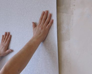 Des mains en train de tenir une bande de papier peint gris clair sur le mur