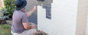 Un homme en chapeau en train de peindre un mur en briques en gris à l’aide d’un rouleau