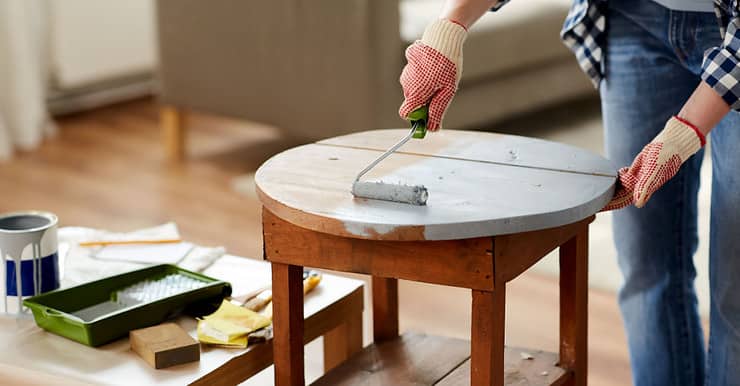 Une femme en gants peint une vieille table en bois à l’aide d’un rouleau de peinture