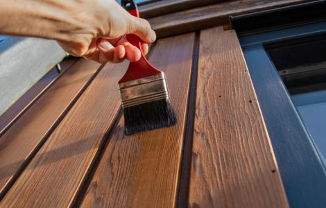 Zoom sur la main d’une personne tenant un pinceau et appliquant du vernis sur une façade extérieure en bois