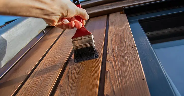 Zoom sur la main d’une personne tenant un pinceau et appliquant du vernis sur une façade extérieure en bois