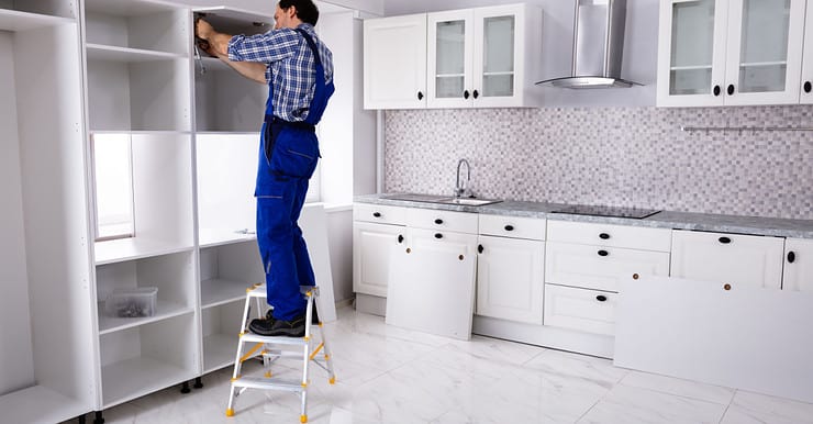 Un homme en combinaison bleue monte sur un escabeau pour monter une étagère dans la cuisine