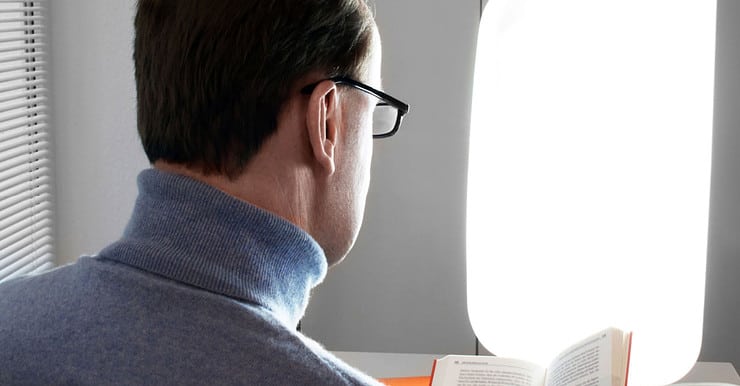Un homme en lunettes près d’une lampe blanche très lumineuse, en train de livre un livre