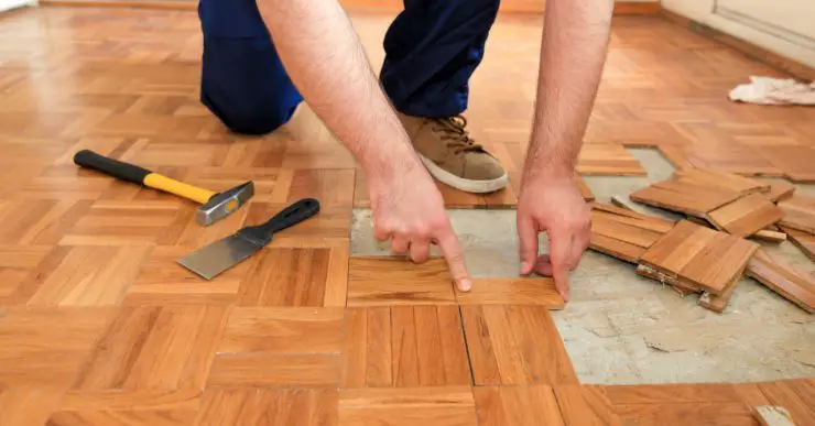 Un homme retire le vieux parquet en bois à l'aide d'un marteau jaune et d'un outil de raclage