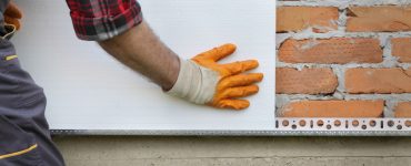 Un homme en gants jaunes installe de l'isolant thermique en polystyrène sur un mur de briques