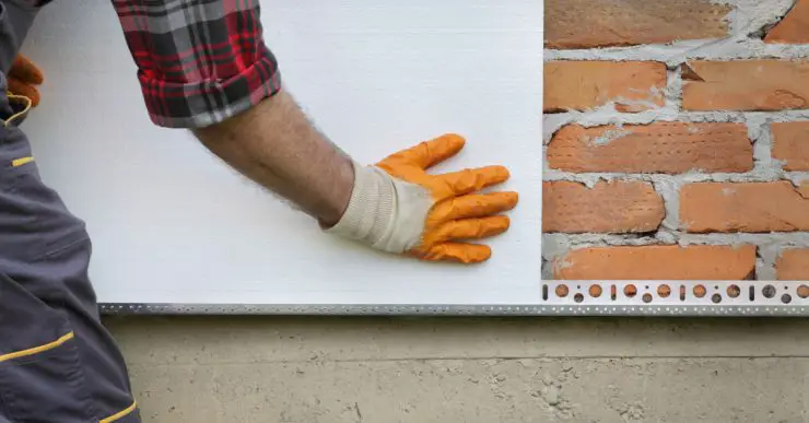 Un homme en gants jaunes installe de l'isolant thermique en polystyrène sur un mur de briques