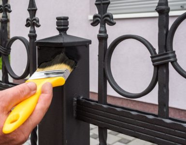 Main d’un homme utilisant un pinceau jaune pour peindre une clôture en fer en noir