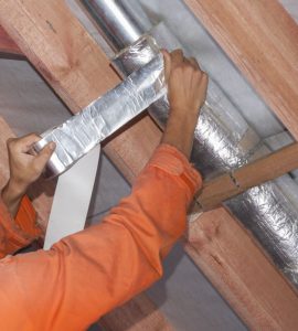 Un ouvrier en tenue de chantier réalise l'isolation d'une ventilation sous le toit