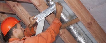 Un ouvrier en tenue de chantier réalise l'isolation d'une ventilation sous le toit