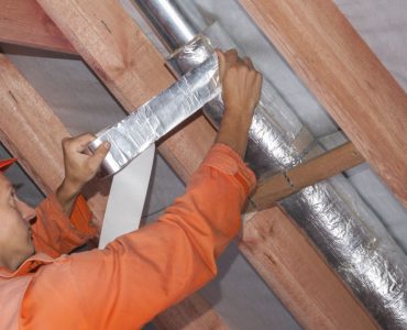 Un ouvrier en tenue de chantier réalise l'isolation d'une ventilation sous le toit