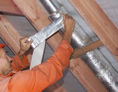 Un ouvrier en tenue de chantier réalise l'isolation d'une ventilation sous le toit