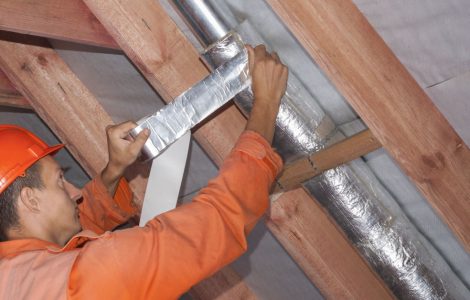 Un ouvrier en tenue de chantier réalise l'isolation d'une ventilation sous le toit