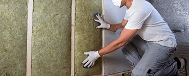 Un homme avec des gants blancs et des lunettes de protection pose un isolant en laine de roche sur un mur