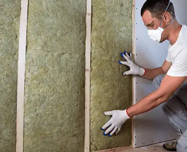 Un homme avec des gants blancs et des lunettes de protection pose un isolant en laine de roche sur un mur