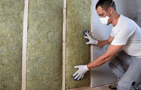 Un homme avec des gants blancs et des lunettes de protection pose un isolant en laine de roche sur un mur