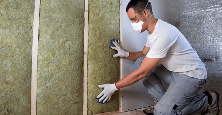 Un homme avec des gants blancs et des lunettes de protection pose un isolant en laine de roche sur un mur