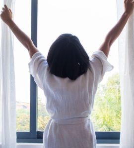 Une femme en robe de chambre ouvrant les rideaux d’une fenêtre