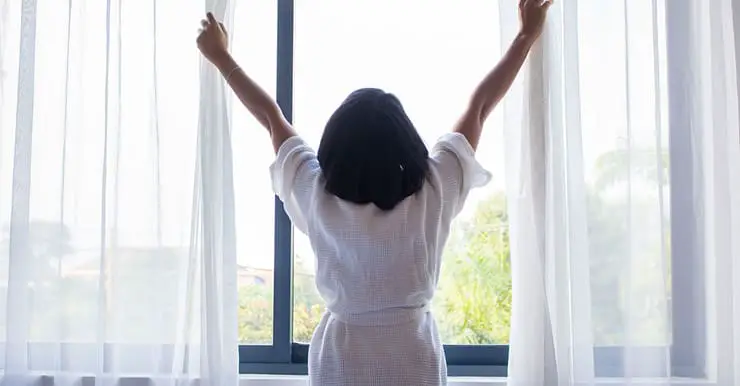 Une femme en robe de chambre ouvrant les rideaux d’une fenêtre