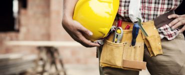 Un homme avec une ceinture porte-outils tient un casque de chantier jaune à la main