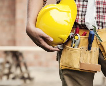 Un homme avec une ceinture porte-outils tient un casque de chantier jaune à la main