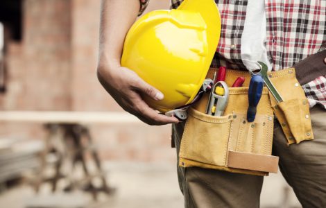 Un homme avec une ceinture porte-outils tient un casque de chantier jaune à la main