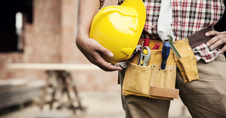 Un homme avec une ceinture porte-outils tient un casque de chantier jaune à la main