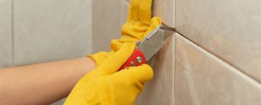 Zoom sur les mains d’une personne en gants jaune utilisant un cutter pour enlever les joints du carrelage mural