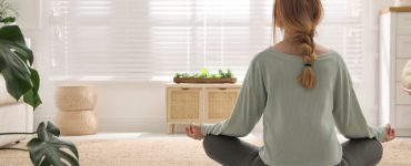 Une femme en position de méditation sur un tapis rond en osier dans son salon