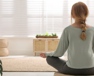Une femme en position de méditation sur un tapis rond en osier dans son salon