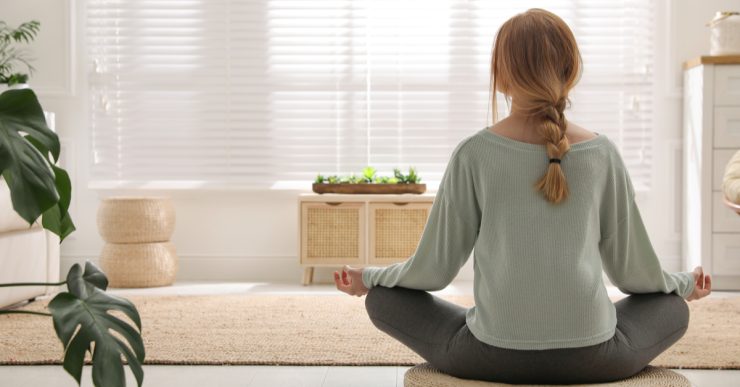 Une femme en position de méditation sur un tapis rond en osier dans son salon