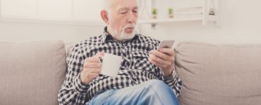 Un homme âgé assis dans un fauteuil, tenant une boisson à la main et regardant son téléphone