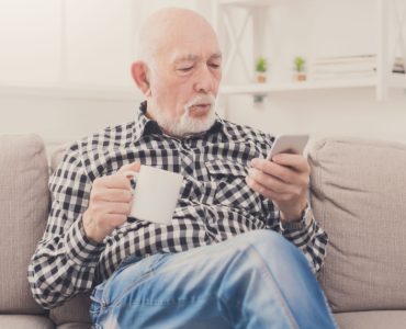 Un homme âgé assis dans un fauteuil, tenant une boisson à la main et regardant son téléphone