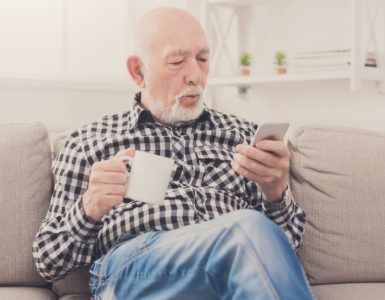 Un homme âgé assis dans un fauteuil, tenant une boisson à la main et regardant son téléphone