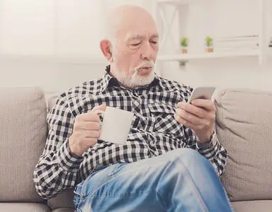 Un homme âgé assis dans un fauteuil, tenant une boisson à la main et regardant son téléphone