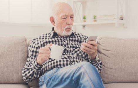 Un homme âgé assis dans un fauteuil, tenant une boisson à la main et regardant son téléphone
