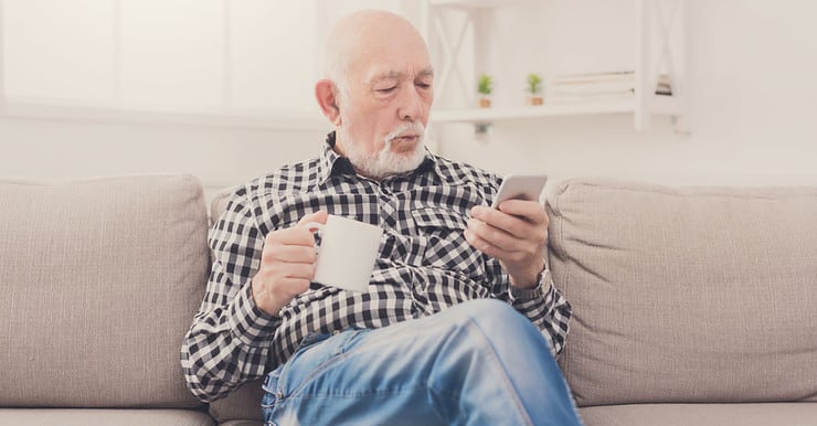 Un homme âgé assis dans un fauteuil, tenant une boisson à la main et regardant son téléphone